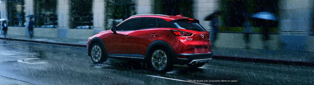 Mazda CX-3 rouge, vu de côté, un peu de dos, roulant sous pluie intense